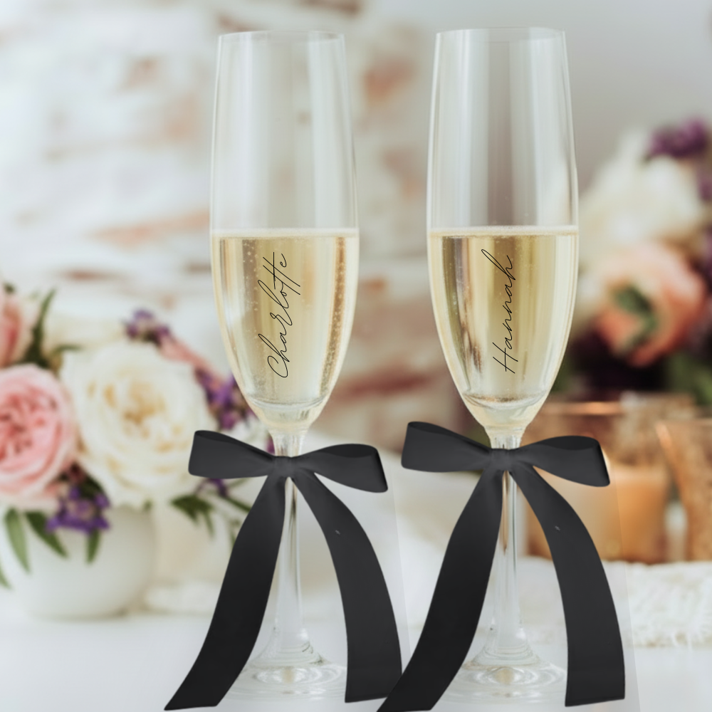 two champagne flutes personalised with minimal script lettering, hand tied black ribbon bows filled with champagne