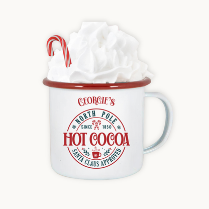 White mug with red rim and 'Georgie's North Pole Hot Cocoa' text, filled with whipped cream and a candy cane.