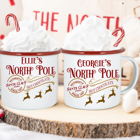 Two mugs labeled 'Ellie's North Pole' and 'Georgie's North Pole' with hot chocolate and whipped cream.
