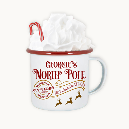 White mug with red rim and 'Georgie's North Pole' text, filled with hot chocolate and whipped cream.