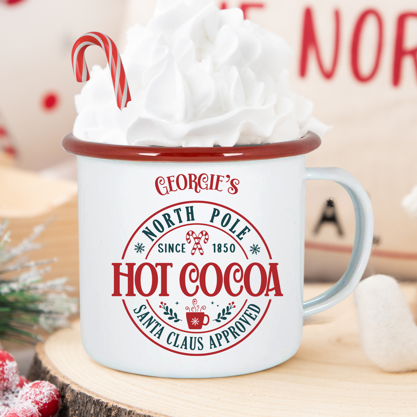 White mug with red rim labeled 'Georgie's North Pole Hot Cocoa' filled with hot chocolate and whipped cream, on a wooden surface.