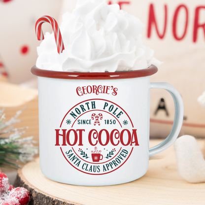 White mug with red rim labeled 'Georgie's North Pole Hot Cocoa' filled with hot chocolate and whipped cream, on a wooden surface.