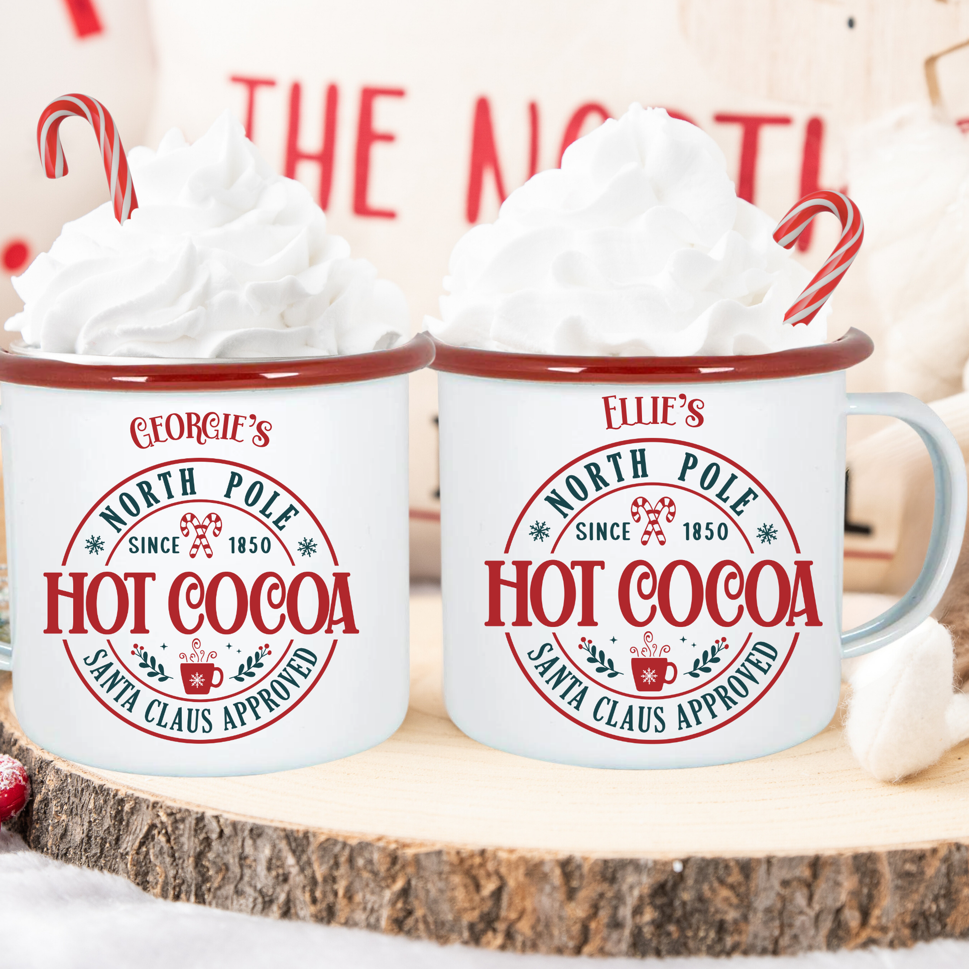 Two mugs labeled 'North Pole Hot Cocoa' with whipped cream and candy canes on a wooden surface.