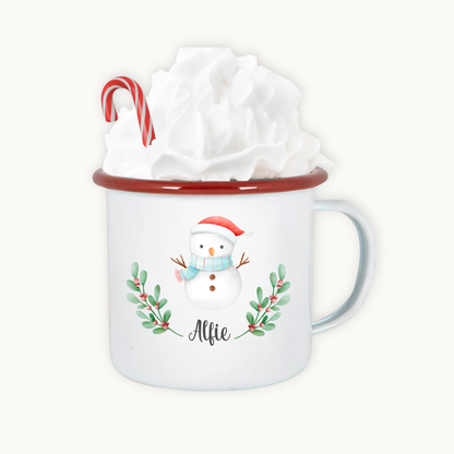 White mug with snowman design and red rim, containing a gift wrapped in white paper.