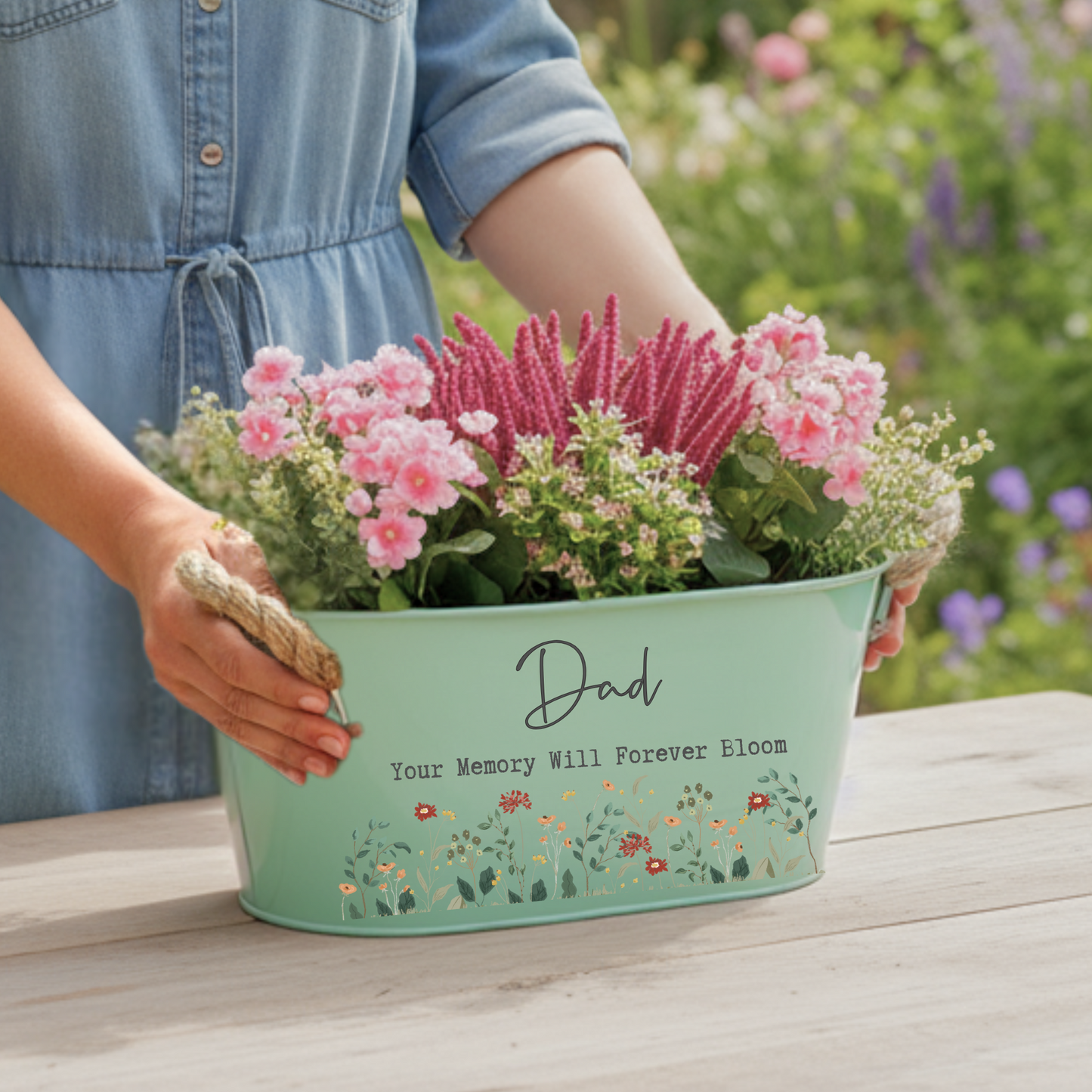 Person holding a planter with flowers and engraved message on a wooden surface.