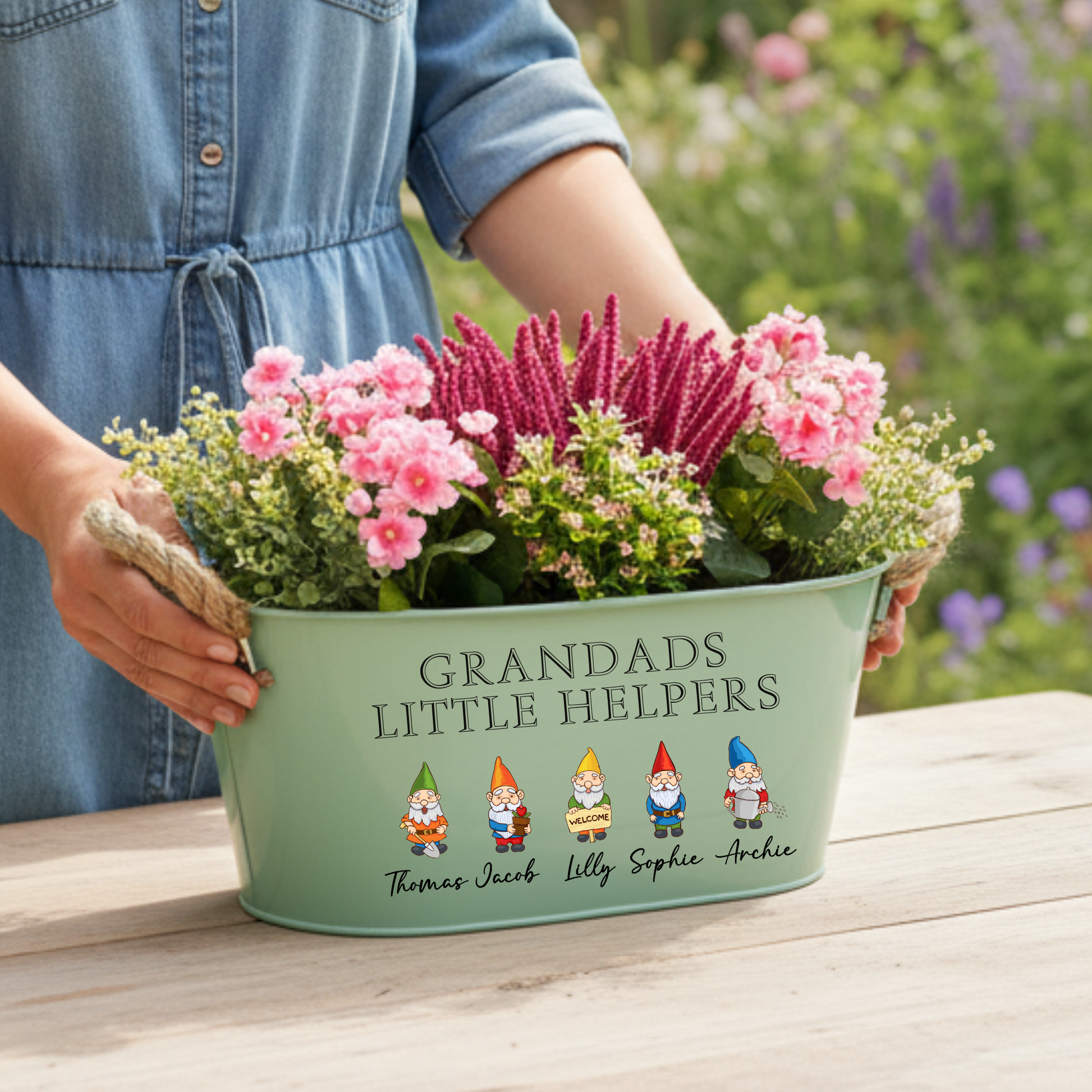 Person holding a green planter with flowers and personalized text in a garden setting
