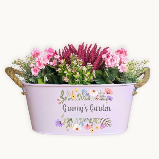 A pink metal garden planter filled with pretty pink flowers and printed with the words Grannys Garden. Surrounded by printed flowers
