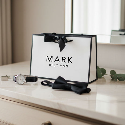 Gift bag labeled 'Mark Best Man' with a bow, watch, and tie on a surface.