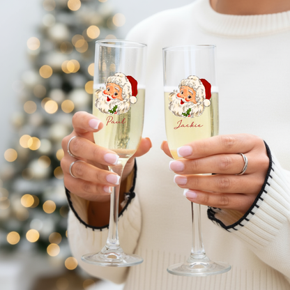 Two people holding champagne flutes with cartoon Santa faces and names against a blurred Christmas tree background.