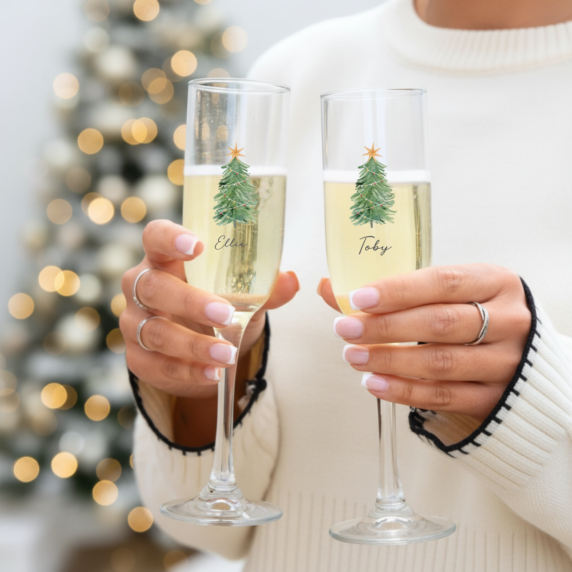 Two people holding champagne glasses with Christmas tree designs and names in front of a blurred festive background.