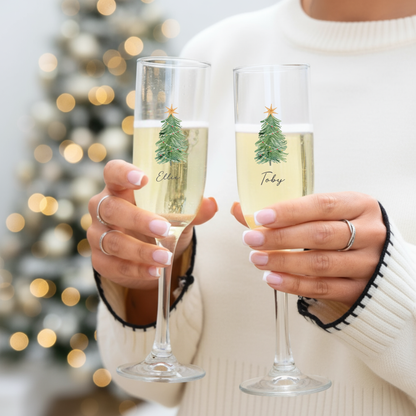 Two people holding champagne glasses with Christmas tree designs and names in front of a blurred festive background.