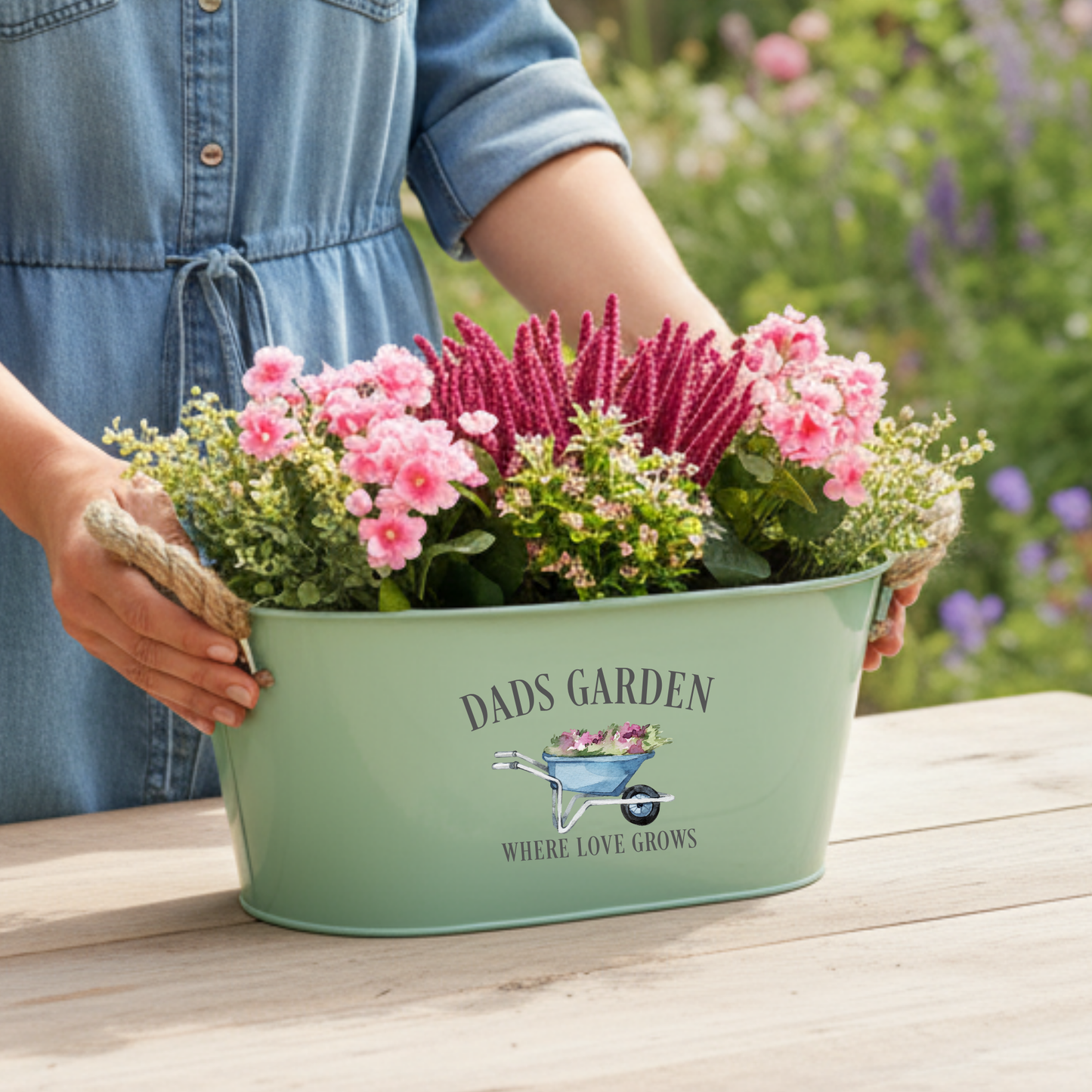 Person holding a green metal tub filled with flowers, featuring 'Dad's Garden' text and design.