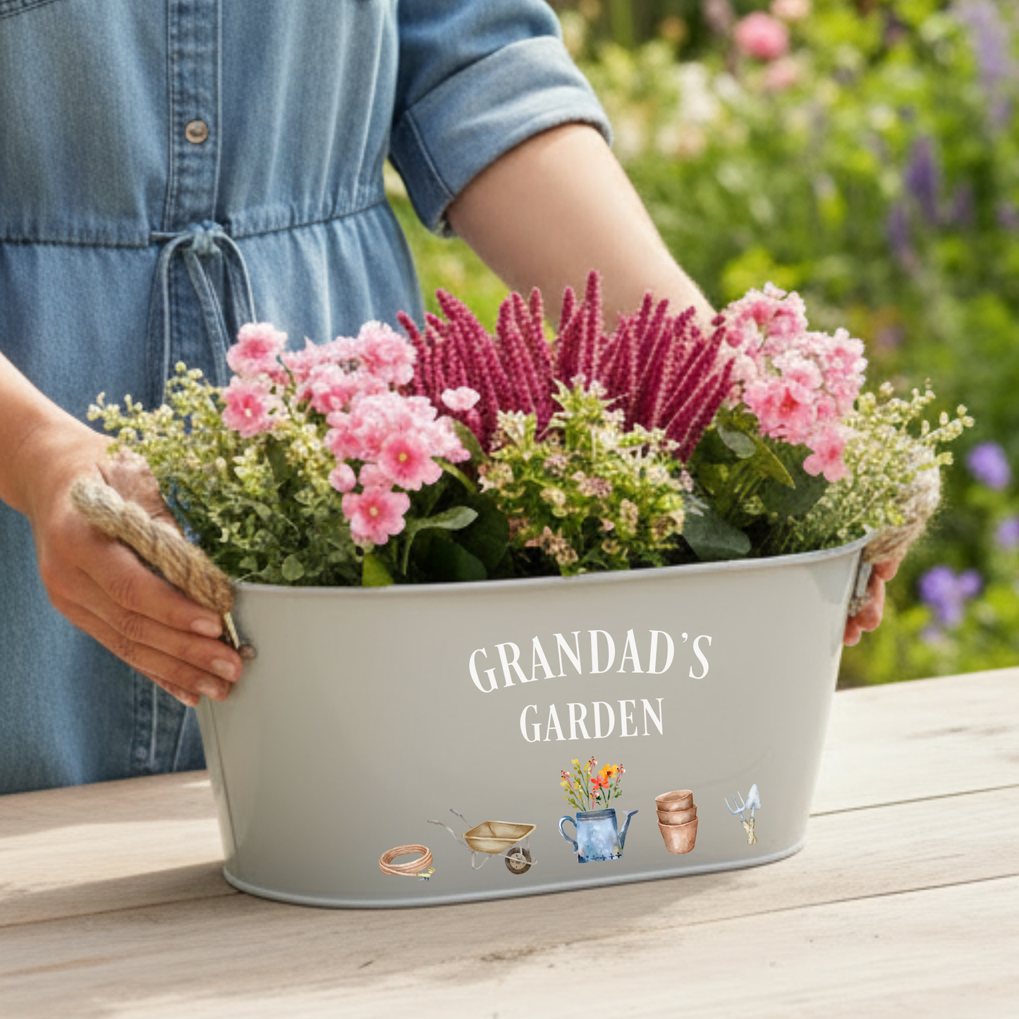 Person holding a planter with flowers labeled 'Grandad's Garden' in a garden setting.