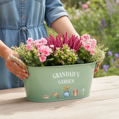 Person holding a green planter with flowers labeled 'Grandad's Garden' in a garden setting.