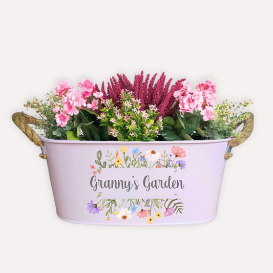 A pink metal garden planter filled with pretty pink flowers and printed with the words Grannys Garden. Surrounded by printed flowers
