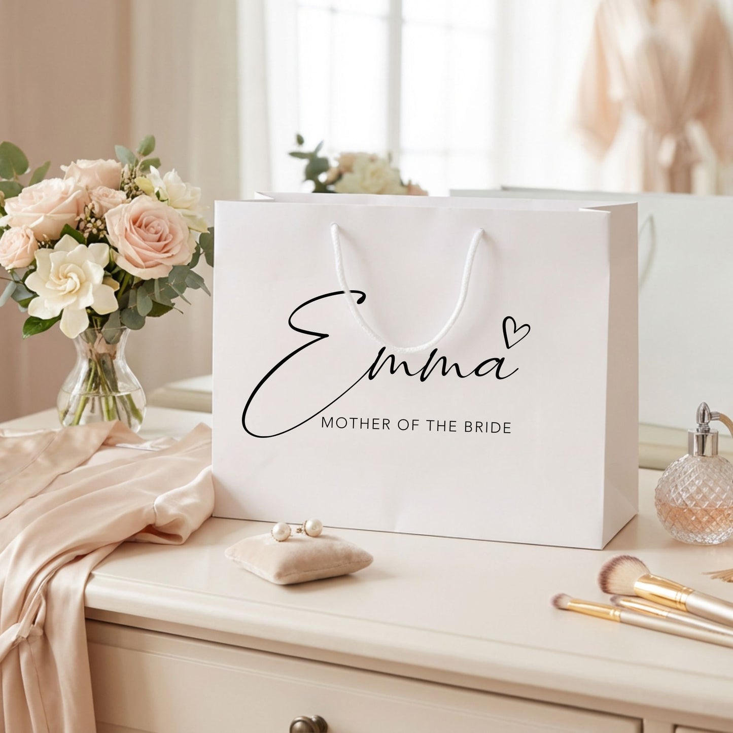 White gift bag with 'Emma Mother of the Bride' text on a table with flowers and makeup items.