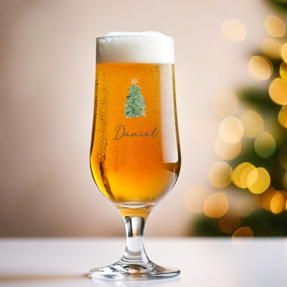 Glass of beer with a Christmas tree design and 'Daniel' text, blurred Christmas tree in the background