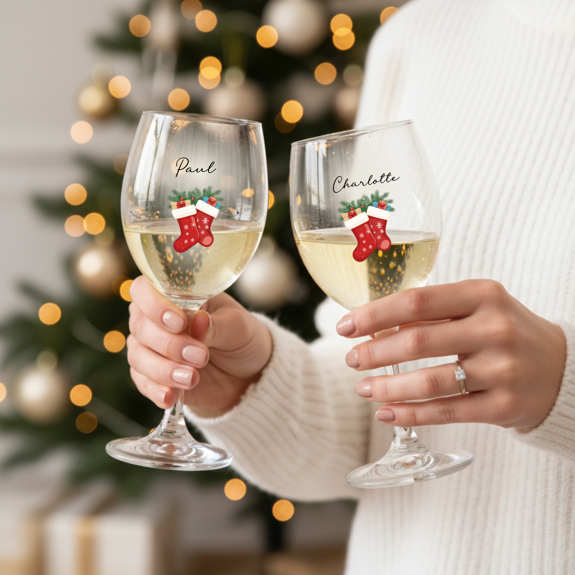 Two wine glasses with personalized designs of Christmas stockings held by a person against a blurred Christmas tree background.