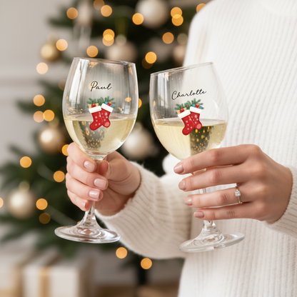 Two wine glasses with personalized designs of Christmas stockings held by a person against a blurred Christmas tree background.