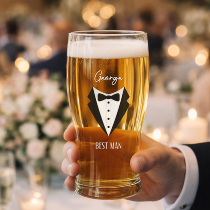 Glass of beer with 'Best Man' and tuxedo design, held by a person in a blurred wedding setting.