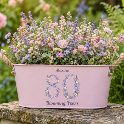 Pink planter with flowers and 'Maxine 80 Blooming Years' text in a garden setting