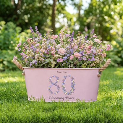 Pink metal planter with flowers and 'Maxine 80 Blooming Years' text on grass