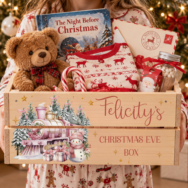 A personalized wooden Christmas Eve gift box filled with holiday treats, a teddy bear, a Christmas book, and a Santa-themed envelope, held by a person wearing matching pajamas in front of a decorated Christmas tree.