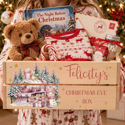 A personalized wooden Christmas Eve gift box filled with holiday treats, a teddy bear, a Christmas book, and a Santa-themed envelope, held by a person wearing matching pajamas in front of a decorated Christmas tree.