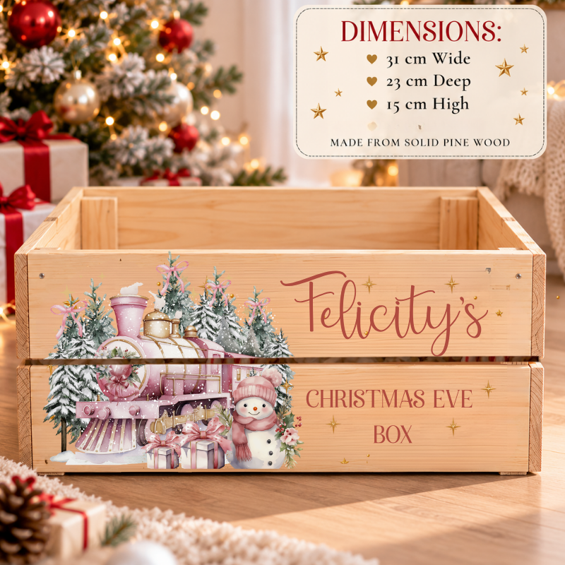 A wooden Christmas Eve box with a festive illustration of a pink train, snowman, and gifts, plus the text 'Felicity's' and 'CHRISTMAS EVE BOX.' A dimension callout in the top right lists the box's measurements, and a blurred Christmas tree and wrapped gifts appear in the background.