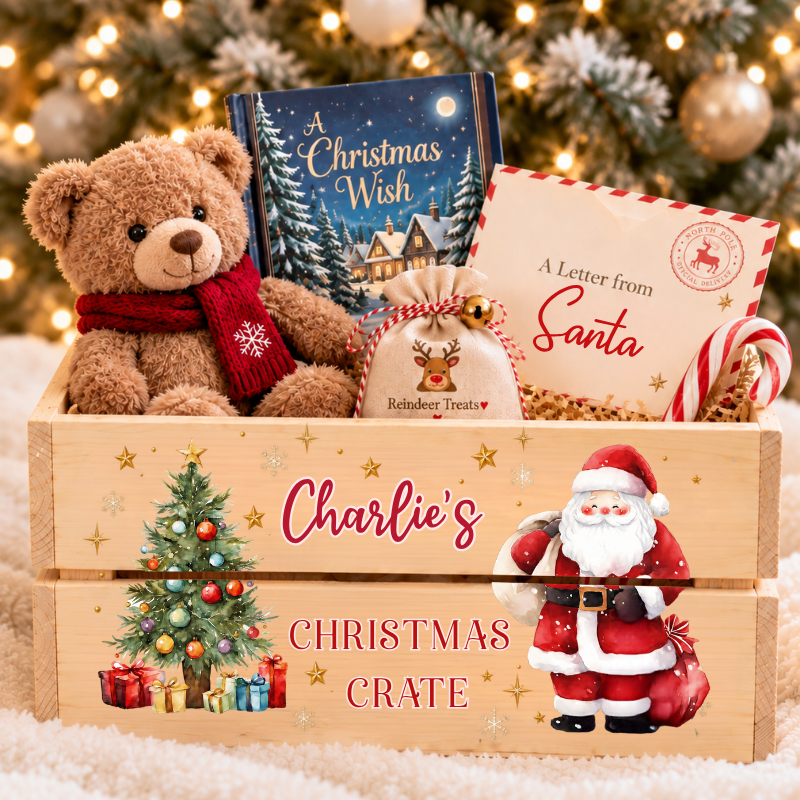 A personalized wooden Christmas crate gift box filled with holiday treats, including a teddy bear, a book titled 'A Christmas Wish', a Santa letter, and candy canes, set against a festive blurred Christmas tree background.