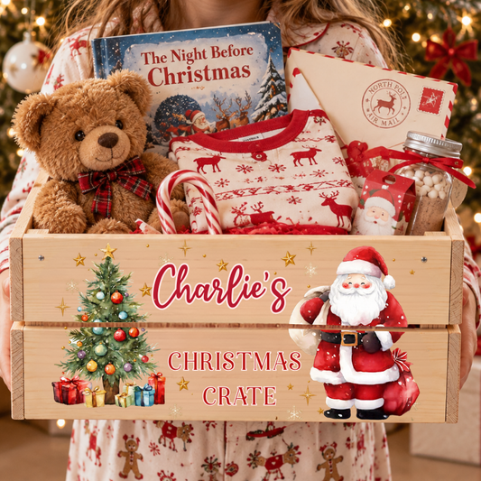 A wooden personalized Christmas crate held by a child wearing matching holiday pajamas. The crate is decorated with a Christmas tree, Santa Claus, and the text 'Charlie's CHRISTMAS CRATE', and contains a teddy bear, a sweater, candy canes, a book, and gift bags.