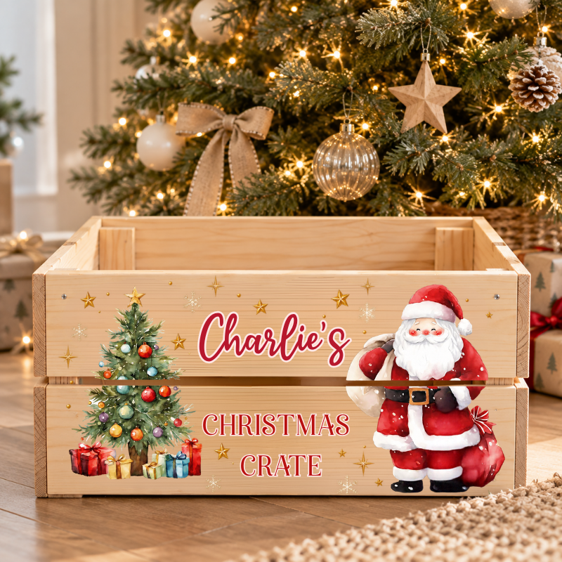 A personalized wooden Christmas storage crate with a printed design featuring a Christmas tree, presents, a Santa Claus figure, and the text 'Charlie's Christmas Crate'. The crate is styled in a festive living room setting in front of a decorated Christmas tree.
