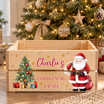 A personalized wooden Christmas storage crate with a printed design featuring a Christmas tree, presents, a Santa Claus figure, and the text 'Charlie's Christmas Crate'. The crate is styled in a festive living room setting in front of a decorated Christmas tree.