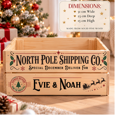 A wooden Christmas-themed shipping crate labeled 'North Pole Shipping Co.' with personalized text 'Evie & Noah,' shown on a wooden floor with a blurred holiday tree and wrapped gifts in the background.