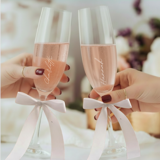 two champgane flutes filled with sparkling rosé prosecco, personalised with white script font and hand tied pale pink ribbon bows on each glass