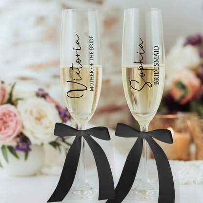 Two champagne flutes with personalized names tied with black ribbons against a blurred floral background.