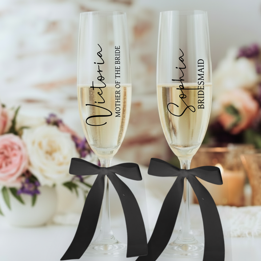 Two champagne flutes with personalized names tied with black ribbons against a blurred floral background.