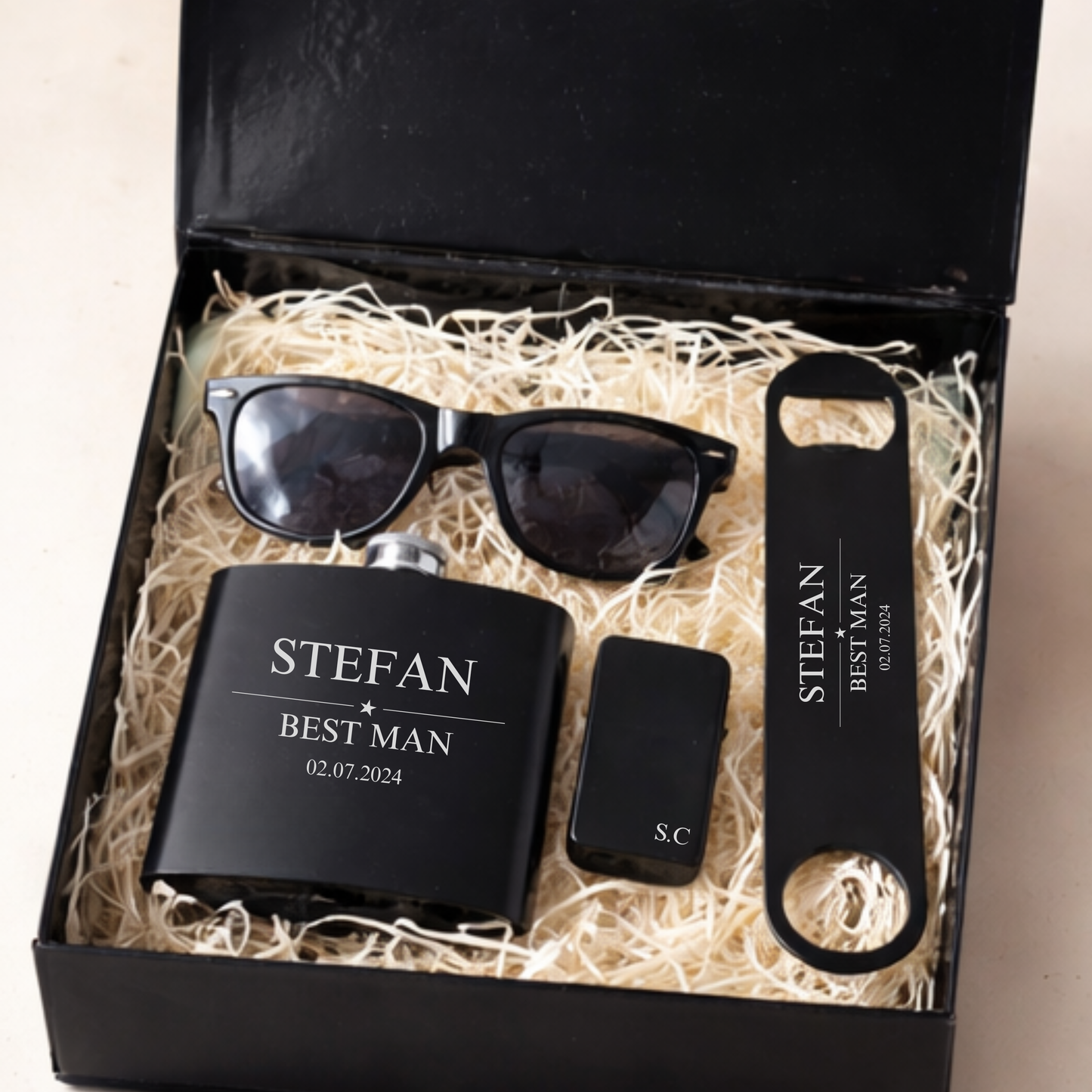 a black gift box filled with gifts for a groomsman or best man, a personalised hip flask, bar blade, sunglasses and zippo lighter, personalised with the name Stefan, Best Man and date. Filled with shreeded wood box filler