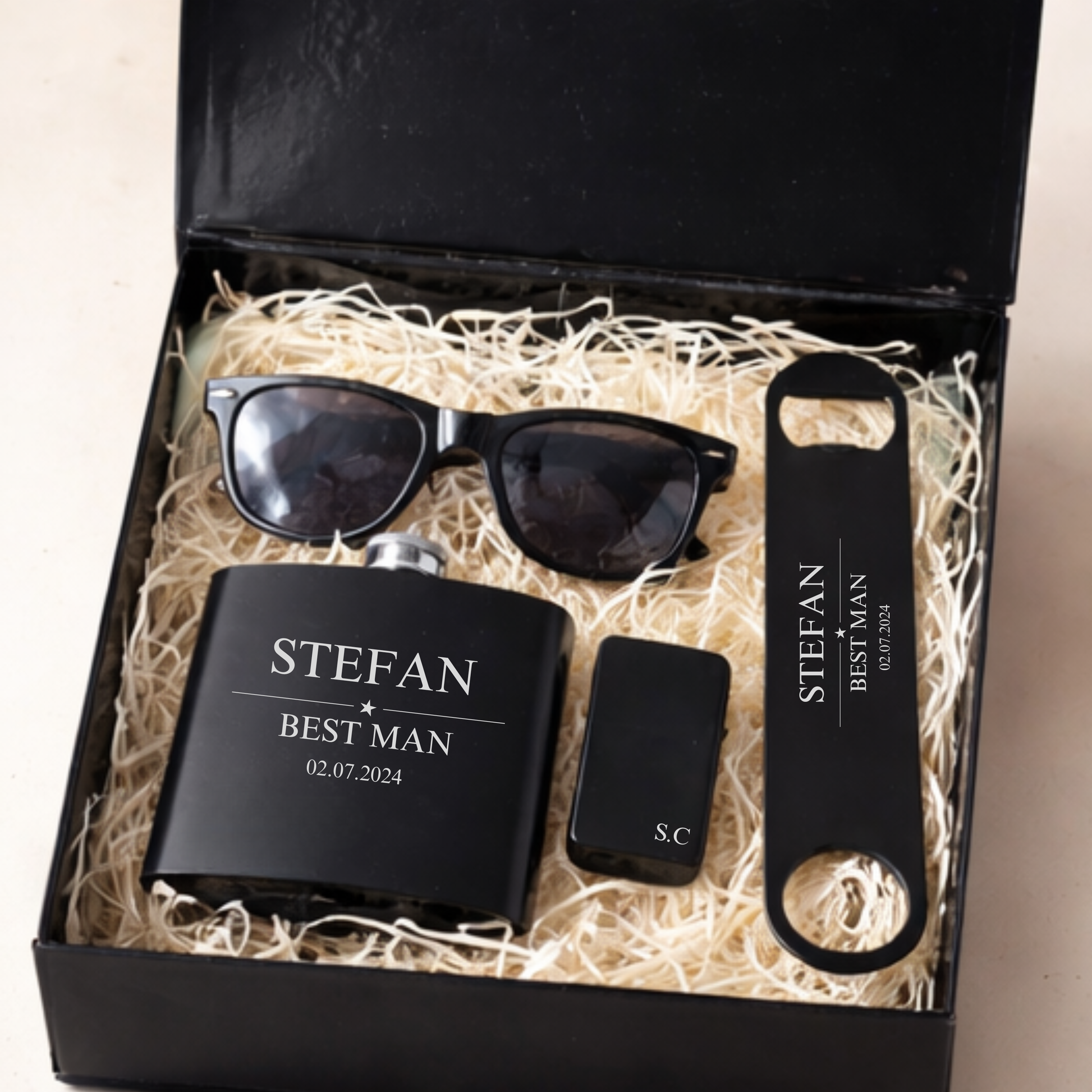 a black gift box filled with gifts for a groomsman or best man, a personalised hip flask, bar blade, sunglasses and zippo lighter, personalised with the name Stefan, Best Man and date. Filled with shreeded wood box filler
